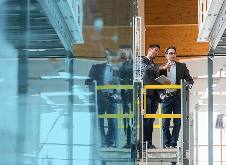 Two business men standing on a riser with yellow safety markings, one holding a tablet, overseeing new construction in a large building