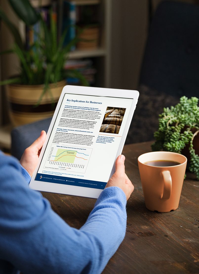 A man in a periwinkle blue sweater is holding a tablet in portrait mode, reading a page entitled, "Key Implications for Business" from a Novo Advisors white paper. An orange coffee mug is to his right on a rustic wooden desk, next to a thriving green jade plant.